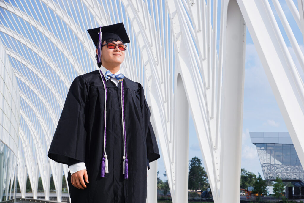 A student in cap and gown stands proudly beneath a series of archways.