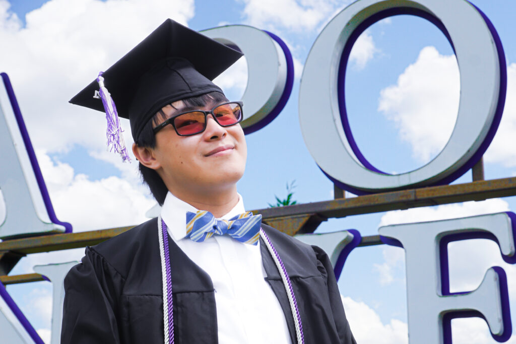 A student in cap and gown smiles and leans his head back in front of a large sign.