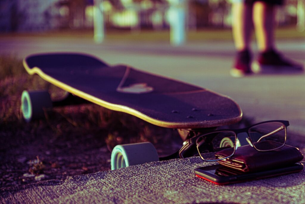 A longboard lying in the grass next to a phone, wallet, and glasses stacked upon each other on a sidewalk.