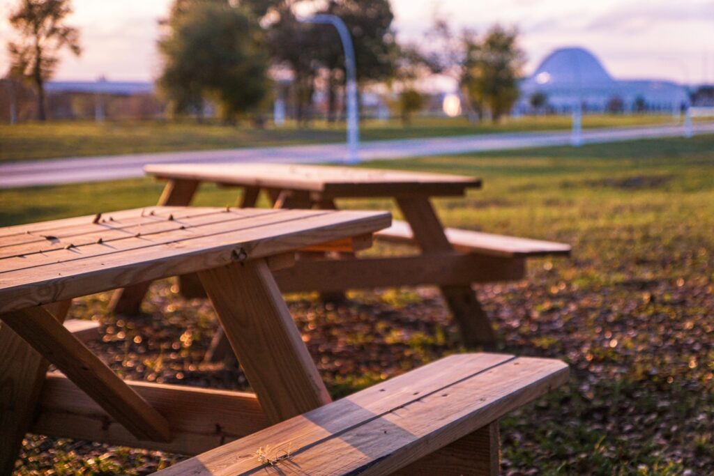 Two picnic tables in a sunlit field with a path and a building in the background.