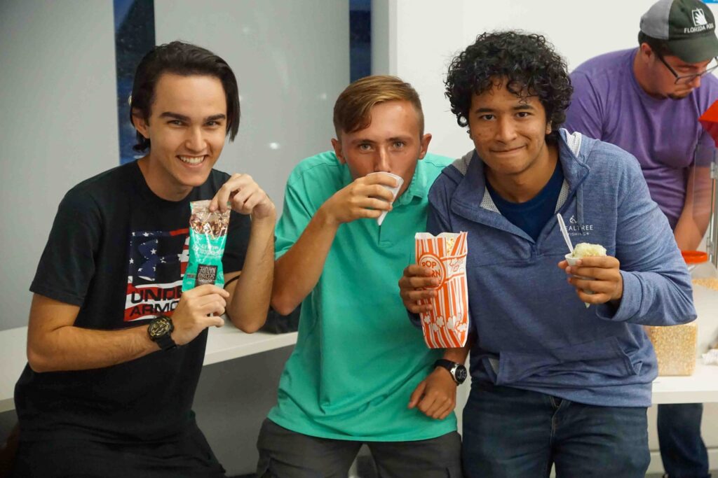 Three boys lean into a photo laughing and holding various snacks.