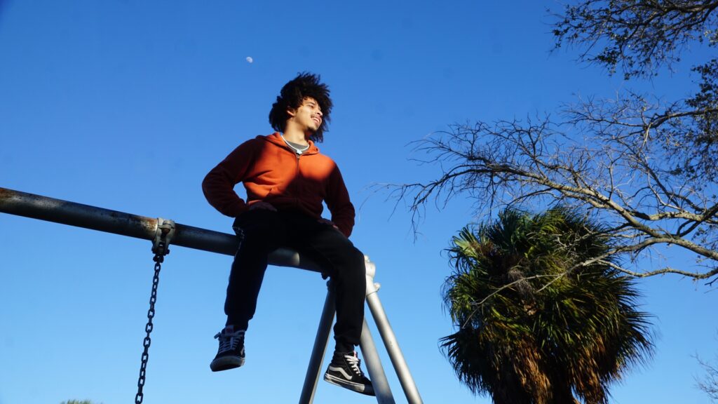 A young man sits on top of a swingset against a blue sky.